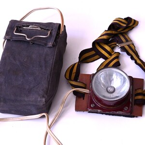 Op de afbeelding: Vintage mijnwerkerslamp set. De set bevat een donkerblauwe canvas accupack met een metalen handvat en een koplamp met een rode metalen behuizing, een heldere lens en een gestreepte band met gele en zwarte strepen.