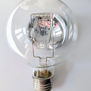 Op de afbeelding: Een heldere, ronde gloeilamp met een zichtbare gloeidraad en een zilveren voet. Het transparante glas biedt zicht op de interne componenten. De voet heeft een schroefontwerp.