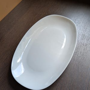 Pode incluir: Uma travessa oval branca com acabamento brilhante. O prato de cerâmica tem bordas suavemente curvas e uma superfície lisa. A travessa está vazia e está sobre uma superfície de madeira marrom escura.