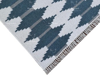 May include: Close-up of a blue and white patterned rug with fringe. The rug has a geometric design with a white background and blue and grey stripes.