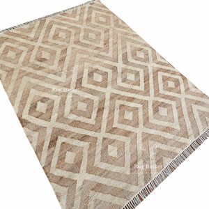 May include: A rectangular rug with a geometric diamond pattern in shades of beige and brown. The rug has a fringe on one end and the text "Rug Basket" is visible. The rug is made of a woven material.