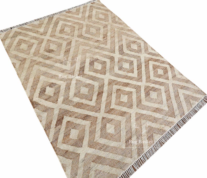 May include: A rectangular rug with a geometric diamond pattern in shades of beige and brown. The rug has a fringe on one end and the text "Rug Basket" is visible. The rug is made of a woven material.