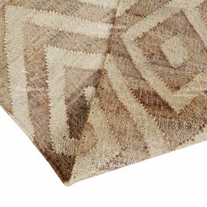 May include: Close-up of a beige and brown geometric patterned rug. The rug features a repeating diamond and stripe design, with varying shades of beige and brown. The rug is made of a woven material.