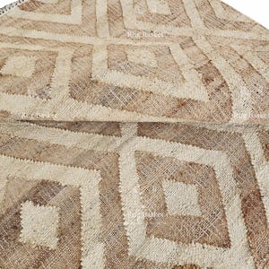 May include: A natural fiber rug with a geometric diamond pattern in shades of beige and brown. The rug is folded over, showcasing the texture and design. The rug is suitable for home decor.