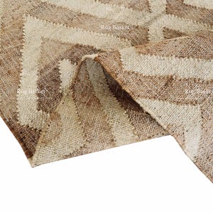 May include: A close-up of a beige and brown rug with a geometric chevron pattern. The rug is made of a woven material and has a textured appearance. The rug is folded over, revealing the underside.
