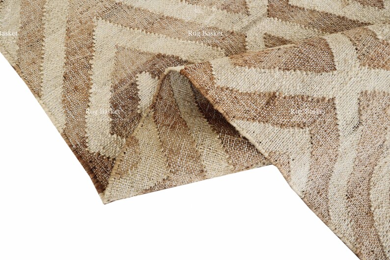 May include: A close-up of a beige and brown rug with a geometric chevron pattern. The rug is made of a woven material and has a textured appearance. The rug is folded over, revealing the underside.