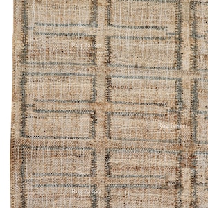 Pode incluir: Close-up de um tapete de juta tecido com um padrão geométrico. O tapete apresenta uma cor bege natural com um design de grade em cinza-azulado suave. A borda inferior tem franjas.