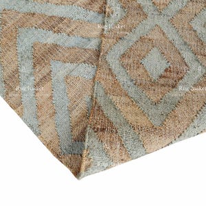 Pu&ograve; includere: Primo piano di un tappeto tessuto con un motivo geometrico. Il tappeto presenta un motivo ripetuto di diamanti e chevron nelle tonalit&agrave; del beige e dell'azzurro. La trama del tappeto &egrave; visibile, suggerendo una costruzione in fibra naturale.