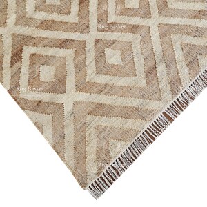 May include: A corner view of a beige and brown geometric patterned rug. The rug features a repeating square design with a fringed edge. The color palette is neutral, with shades of beige and brown.