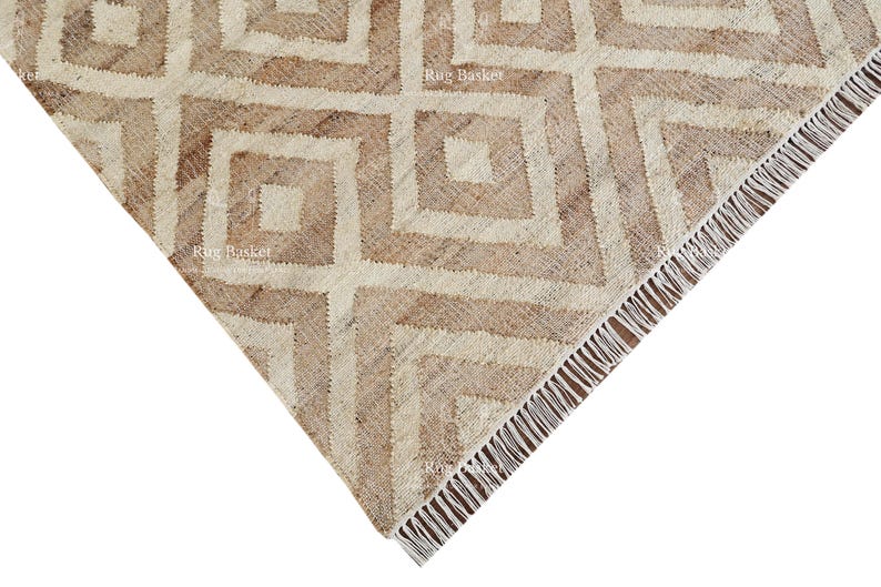 May include: A corner view of a beige and brown geometric patterned rug. The rug features a repeating square design with a fringed edge. The color palette is neutral, with shades of beige and brown.