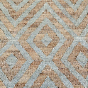 Pu&ograve; includere: Primo piano di un tappeto tessuto con un motivo geometrico. Il tappeto presenta un design ripetitivo di diamanti e zig-zag nei toni del beige, azzurro e marrone. La trama del tappeto &egrave; visibile, suggerendo un materiale in fibra naturale.