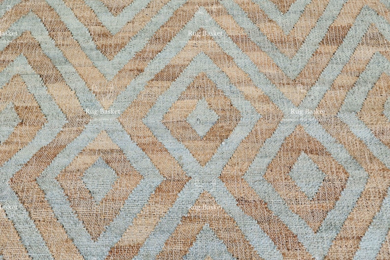 Pu&ograve; includere: Primo piano di un tappeto tessuto con un motivo geometrico. Il tappeto presenta un design ripetitivo di diamanti e zig-zag nei toni del beige, azzurro e marrone. La trama del tappeto &egrave; visibile, suggerendo un materiale in fibra naturale.