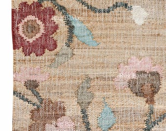 May include: Close-up of a beige jute rug with an embroidered floral design. The rug features flowers in shades of red, pink, and brown, with blue and green leaves. The rug has a fringed edge.