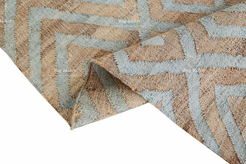 Pu&ograve; includere: Un tappeto tessuto con un motivo geometrico. Il tappeto presenta fasce alternate di blu chiaro e beige, creando un design a zig-zag. Il tappeto &egrave; realizzato in un materiale strutturato.
