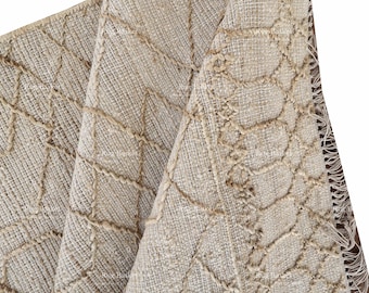 May include: Beige woven rug with a geometric pattern. The rug features a textured surface with a repeating design of interconnected shapes. The edges of the rug have a fringed detail. The rug is folded over, showing the underside.