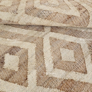 May include: Close-up of a woven rug with a geometric diamond pattern. The rug features a repeating design of diamonds in shades of beige and brown, creating a textured surface. The rug is made of natural fibers.