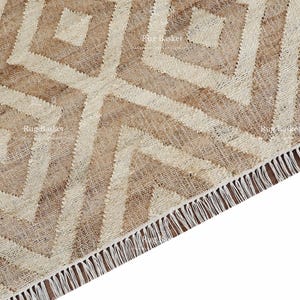 May include: Close-up of a beige and cream-colored rug with a geometric diamond pattern. The rug has a fringed edge and is made of a woven material. The design features repeating diamond shapes.