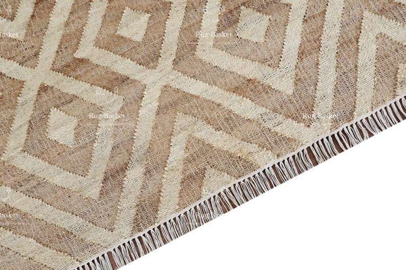 May include: Close-up of a beige and cream-colored rug with a geometric diamond pattern. The rug has a fringed edge and is made of a woven material. The design features repeating diamond shapes.