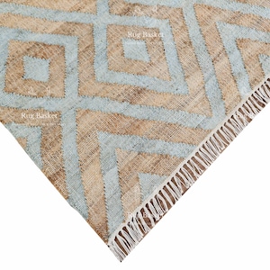 Pu&ograve; includere: Primo piano di un tappeto geometrico con un motivo a rombi. Il tappeto presenta una combinazione di colori azzurro e beige. Il bordo del tappeto ha un dettaglio a frange. Il tappeto &egrave; realizzato in un materiale tessuto.