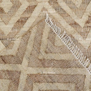 May include: A close-up of a beige and brown geometric patterned rug. The rug features a repeating diamond and zigzag design. The edge of the rug is visible, showing a fringe detail.