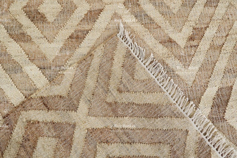 May include: A close-up of a beige and brown geometric patterned rug. The rug features a repeating diamond and zigzag design. The edge of the rug is visible, showing a fringe detail.