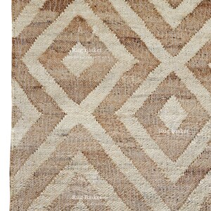 May include: Close-up of a natural fiber rug with a geometric diamond pattern. The rug features alternating shades of beige and brown, creating a textured, woven design. The edge has a fringe detail.