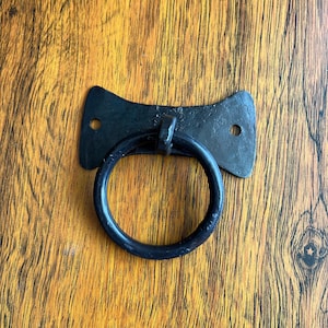 以下が含まれることがあります： 丸みを帯びた滑らかなリングが付いた黒い金属製のリングプルハンドル。ハンドルは、丸みを帯びたエッジが付いた平らな長方形の金属プレートに取り付けられています。