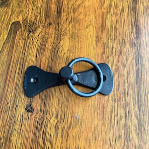 Op de afbeelding: Zwarte metalen deurgreep met een ringtrekker. De greep is bevestigd aan een houten oppervlak.