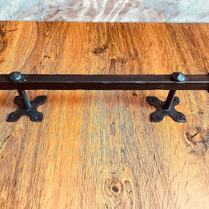 Op de afbeelding: Zwarte metalen handdoekhouder met decoratieve eindkappen. De houder is gemonteerd op een houten oppervlak.