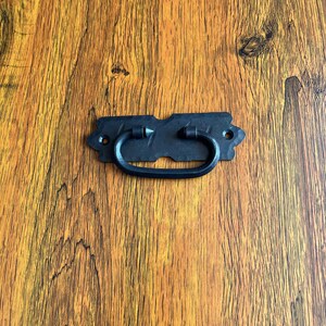 Könnte beinhalten: Schwarzer Metall-Schubladengriff mit schlichtem, elegantem Design. Der Griff hat eine rechteckige Form mit abgerundeten Kanten und einem leicht gebogenen Griff.