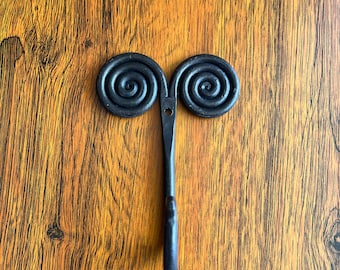 May include: Black metal hook with a double spiral design. The hook is mounted on a wooden surface.