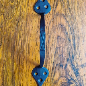 May include: A black, wrought iron door pull with a decorative design. The handle features a central bar with an X-shaped detail, connecting two heart-shaped ends, each with three circular holes. The hardware is set against a warm, wood-grain background.