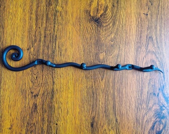 May include: A black metal coat rack with a decorative swirl at one end and five hooks. The rack is mounted on a wooden wall.