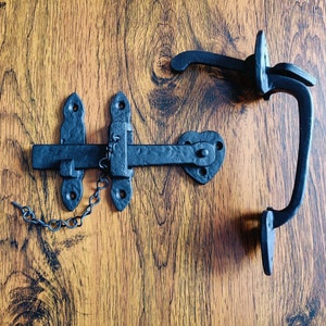 May include: Black metal gate latch with a chain and a separate handle. The latch has a rectangular bar with a hook on one end and a square plate on the other. The handle is a curved piece of metal with a loop on the end.