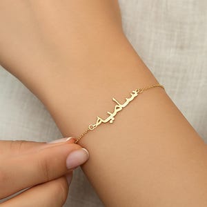 May include: A gold-colored bracelet with the Arabic name "سليميه" is displayed on a wrist. The delicate chain and nameplate are visible against the skin. The bracelet is a personalized piece of jewelry.