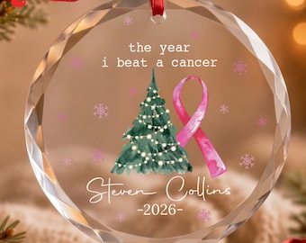 Könnte beinhalten: Ein runder, klarer, facettierter Glasanhänger mit einem aquarellierten grünen Weihnachtsbaum, einer rosa Schleife und dem Text: the year i beat a cancer, Steven Collins, und 2026. Er hängt an einem roten Band vor einem unscharfen Hintergrund.