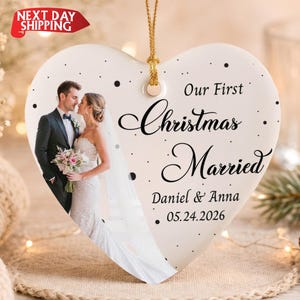 May include: A heart-shaped white ceramic ornament featuring a photograph of a couple in wedding attire. The text reads Our First Christmas Married, Daniel & Anna, and 05.24.2026. It includes a gold hanging string and black polka dot accents.