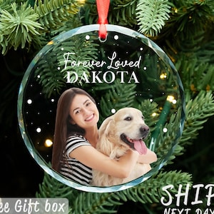 May include: A clear, round Christmas ornament with a photo of a person and a dog, the text "Forever Loved. DAKOTA" is printed on the front. The ornament hangs from a red ribbon against a backdrop of green pine needles.
