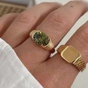 May include: Two gold rings. One ring features a large, oval, olive-green gemstone set in a ribbed gold band. The second ring is a gold signet ring with a rectangular face and a textured band.