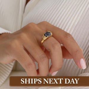 May include: A gold ring with a large, dark gemstone. The ring has a detailed band with a ribbed design. The gemstone is oval-shaped and set in a gold bezel. The ring is on a person's finger.