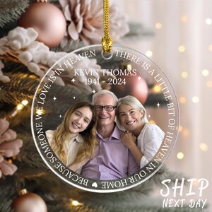 May include: A clear glass Christmas ornament with a photo of a family of three. The ornament has the text "THERE IS A LITTLE BIT OF HEAVEN IN OUR HOME BECAUSE SOMEONE WE LOVE IS IN HEAVEN" and "KEVIN THOMAS 1941-2024".