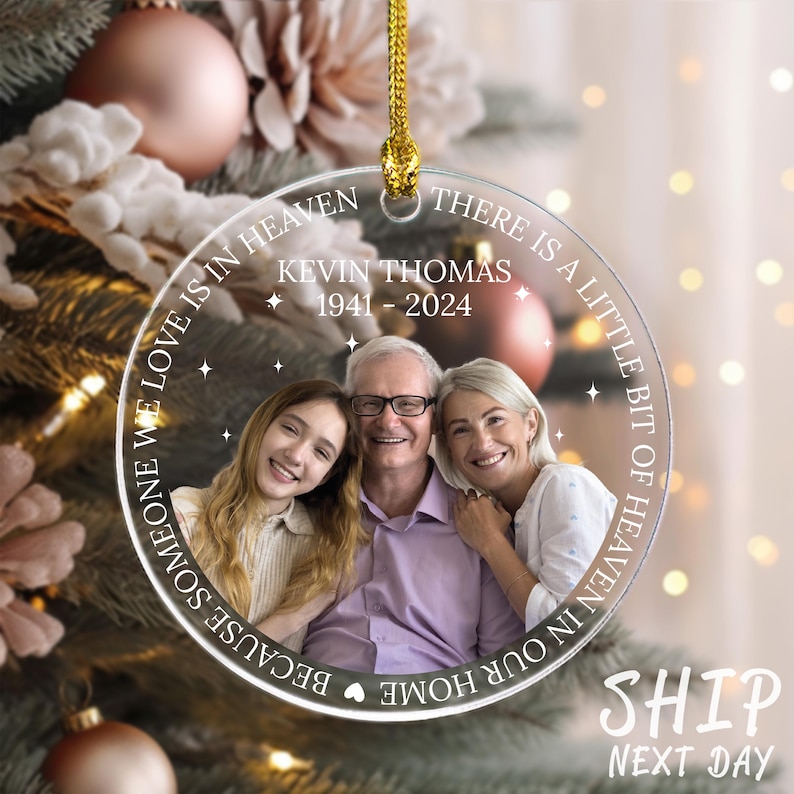 May include: A clear glass Christmas ornament with a photo of a family of three. The ornament has the text "THERE IS A LITTLE BIT OF HEAVEN IN OUR HOME BECAUSE SOMEONE WE LOVE IS IN HEAVEN" and "KEVIN THOMAS 1941-2024".