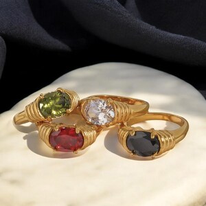 May include: Four gold-colored rings with oval-shaped gemstones in green, clear, red, and black. The rings have a ribbed design around the gemstone settings. The rings are displayed on a white surface with a dark background.
