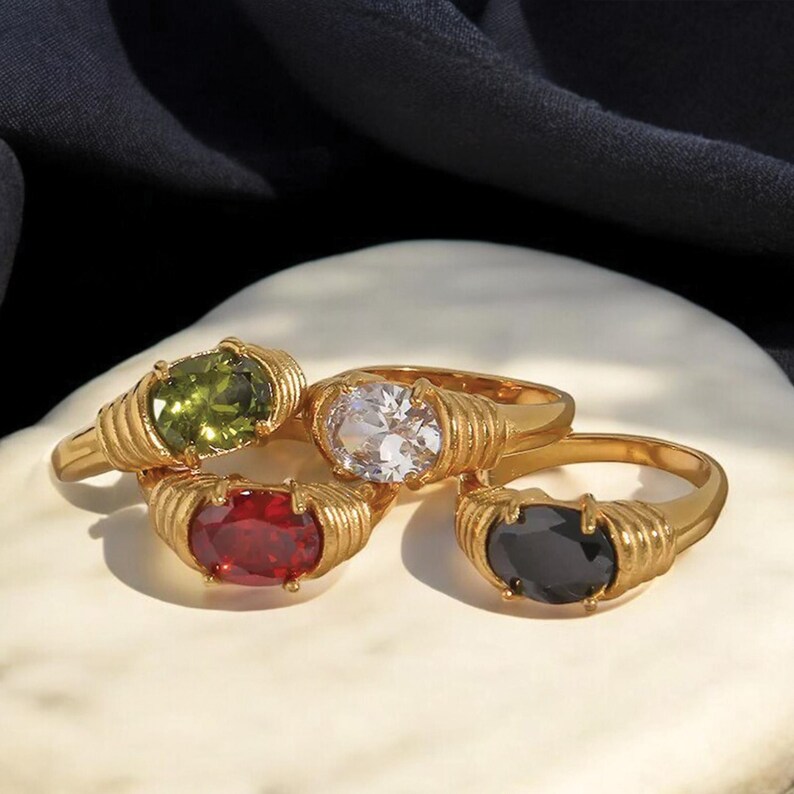 May include: Four gold-colored rings with oval-shaped gemstones in green, clear, red, and black. The rings have a ribbed design around the gemstone settings. The rings are displayed on a white surface with a dark background.