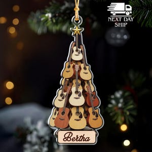 May include: A Christmas tree ornament shaped like a tree, composed of multiple acoustic guitars in various shades of brown. The ornament has a gold star and a wooden plaque with the name "Bertha".