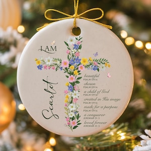 May include: Round ceramic ornament with a floral cross design. The ornament features the name "Scarlet" and the words "I AM". Additional text includes Bible verses about being beautiful, chosen, and loved. A gold string is attached for hanging.