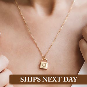 May include: A gold necklace with a padlock pendant featuring the letter "A" engraved on it. The necklace is worn by a person with their chest and shoulders visible.
