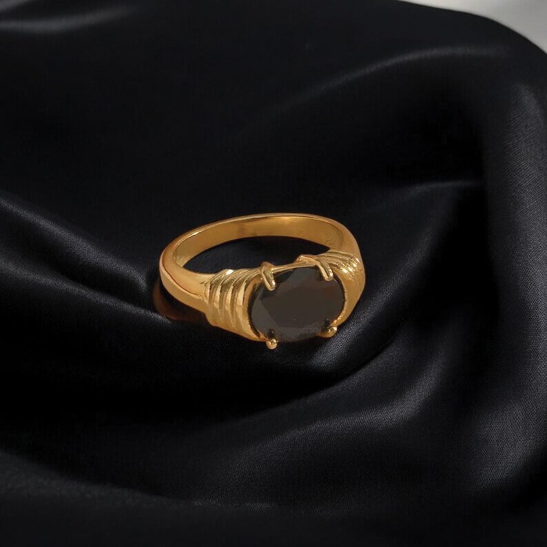 May include: A gold-colored ring with a large, oval-shaped black stone. The ring has a textured design on the sides and is set against a black background.