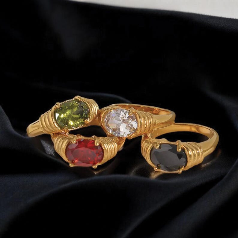 May include: Four gold-colored rings with oval-shaped gemstones in green, clear, red, and black. The rings have a ribbed design around the gemstone settings. The rings are displayed on a black surface.