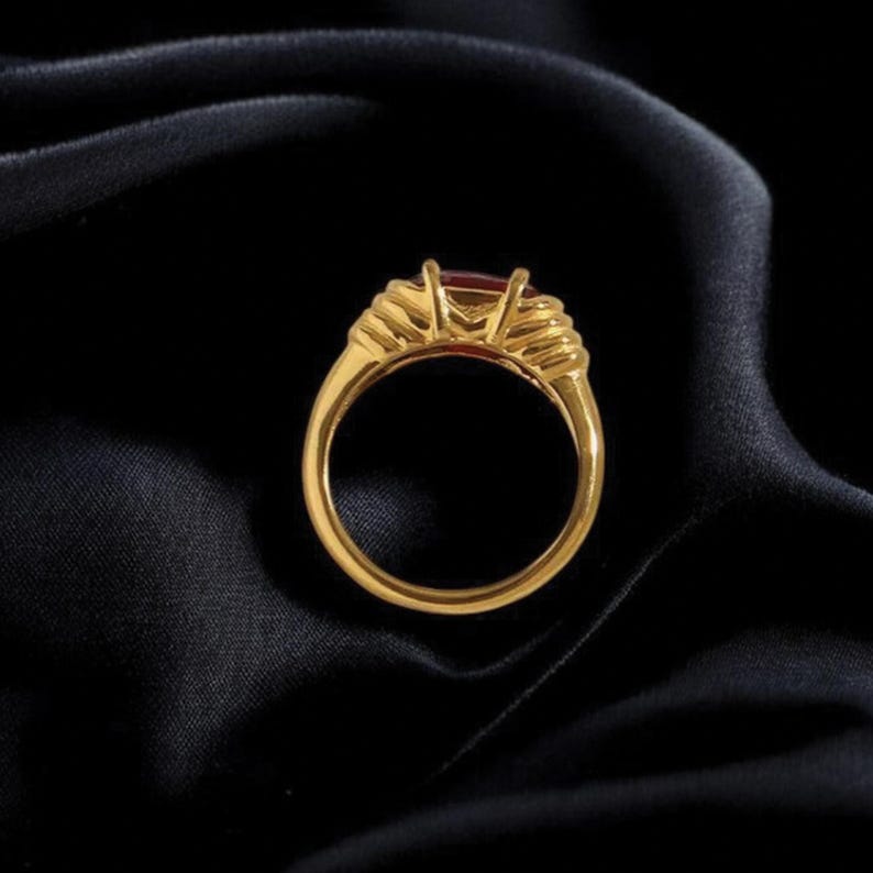 May include: A gold ring with a rectangular red gemstone. The ring has a detailed design on the sides of the gemstone setting. The ring is set against a black background.